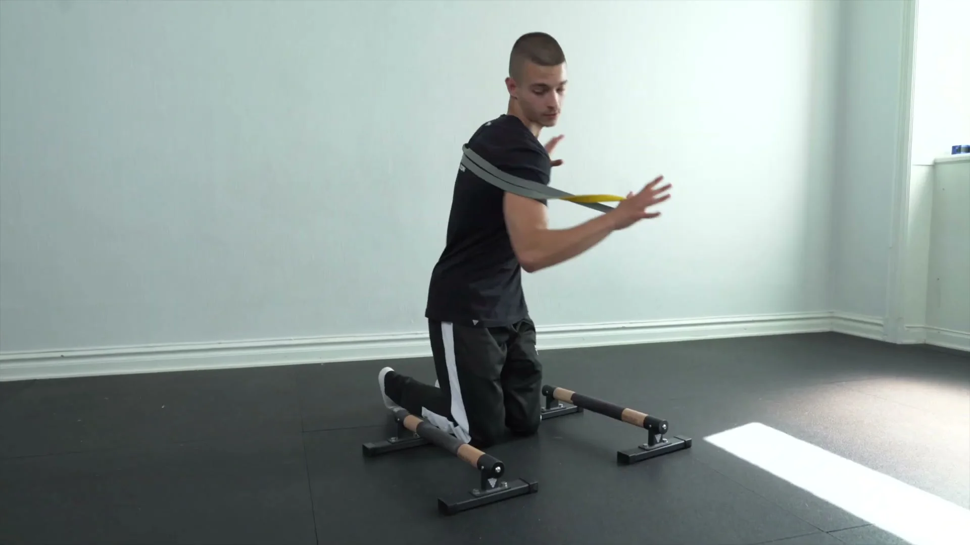 Daniel Hristov standing with a resistance band looped across his back and held in both hands, demonstrating the band setup before getting into the lean push-up position on parallettes for tip two of the explosive planche push-up tutorial