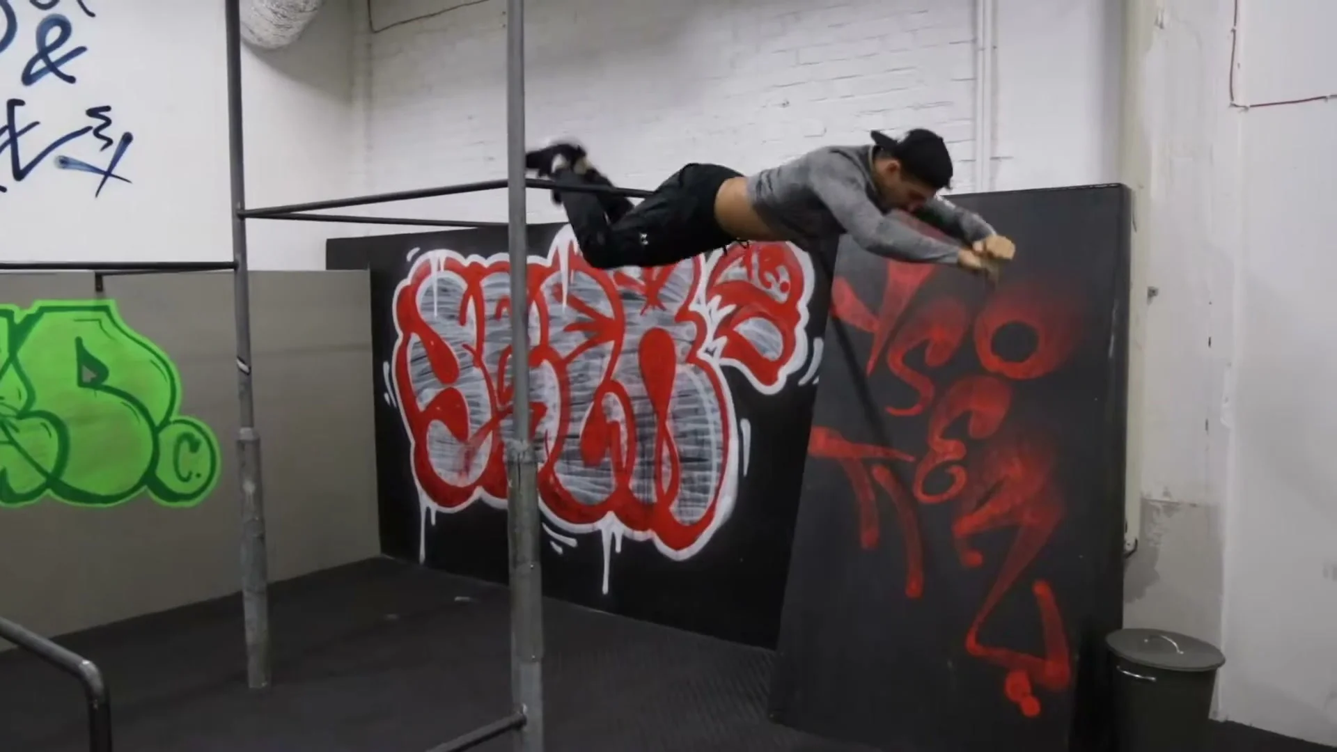 Daniel Flefil performing a knee gainer on a bar in a graffiti-decorated gym, body horizontal in flight after releasing the bar by the knees