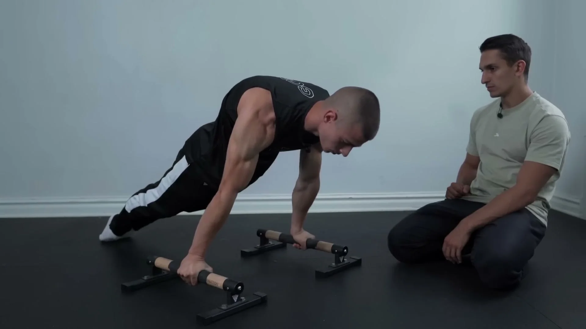 Daniel Hristov performing a lean push-up on parallettes: body in the forward lean position executing a push-up, with Daniel Flefil watching from the side to check form